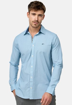 Camisa de botones de color azul claro con un patrón de lunares texturizados. Presenta un cuello en contraste de un oscuro navy y un logo bordado en el pecho.