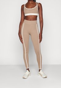 Beige sport-bh och tights i stretchigt material. Vita sidostråk på tightsen, med en logotyp på benet. Ljust färgade träningsskor.