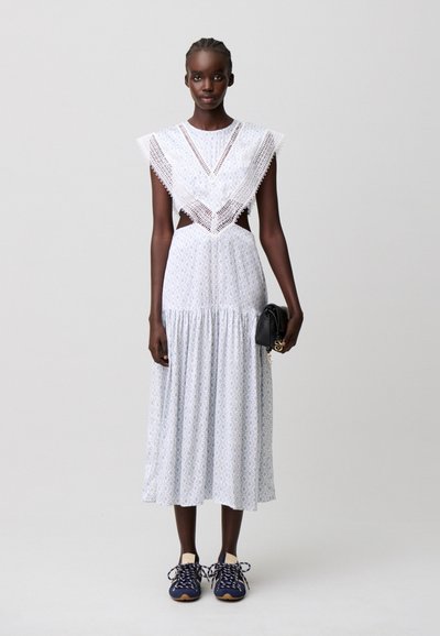 Donna che indossa un vestito midi bianco con dettagli in pizzo, tiene una pochette nera e indossa sneaker blu scuro, in piedi davanti a uno sfondo semplice.