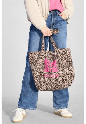 Osoba v béžové bundě, růžovém topu, modrých džínách a bílých teniskách, držící kabelku s leopardím vzorem a růžovým logem "Studio".