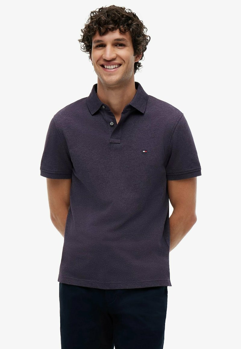 Homme aux cheveux bouclés souriant, portant un polo violet foncé à manches courtes et un pantalon foncé, debout les mains dans le dos.