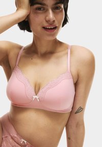 Ljusrosa bh med spetsdetaljer längs halsringningen och centralt, utrustad med justerbara axelband och en liten rosett i mitten fram.