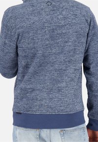 Widok z tyłu osoby w niebieskim, teksturowanym, długim swetrze z ribbanym dołem oraz jasnoniebieskich dżinsach.