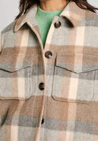 Chaqueta de lana a cuadros en tonos beige, gris y crema, llevada sobre un suéter de punto verde claro.