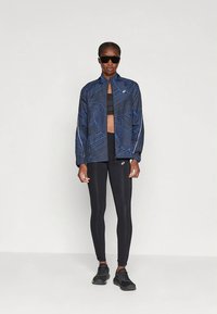 Veste de sport bleu marine à motif géométrique, associée à un soutien-gorge de sport noir et des leggings. Des baskets noires complètent la tenue.