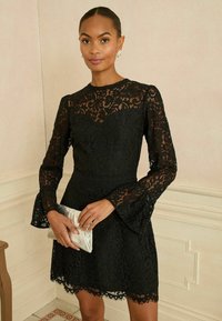 A fekete csipkebal dressz hosszú harangujjakkal, testhez álló felsőrésszel és fodros alsó szegéllyel rendelkezik. A modell egy ezüst színű clutch táskát tart.