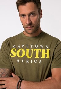 Olivově zelené tričko s výrazným bílým a žlutým textem "CAPE TOWN SOUTH AFRICA" s jemnou texturou. Krátké rukávy, kulatý výstřih.