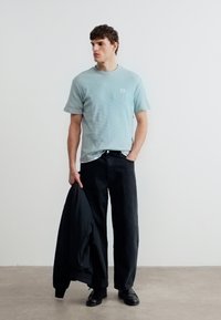 Νέος άνδρας που στέκεται, φοράει ανοιχτό μπλε κοντομάνικο πουκάμισο και μαύρο παντελόνι, κρατώντας ένα μαύρο σακάκι στο ένα χέρι, απλό φωτεινό φόντο.