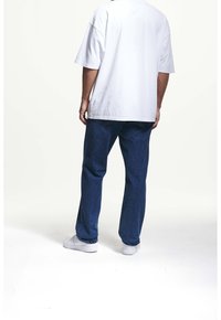 Biały bawełniany T-shirt o luźnym kroju, zestawiony z ciemnoniebieskimi dżinsami. Model nosi białe trampki. Prosty, casualowy strój widziany od tyłu.