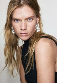 Číre akrylové visiace náušnice s geometrickým dizajnom, nosené osobou s dlhými, vlnitými blond vlasmi, oblečenou v čiernom topánkovom topu s vysokým golierom.