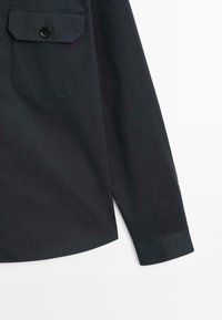 Dark shirt with long sleeves, made of smooth fabric. Features a patch pocket with a button, and rounded hemline.