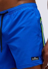 Pantaloni da bagno blu realizzati in materiale leggero, con vita regolabile tramite cordino, dettagli laterali in verde e una piccola etichetta nera del marchio.