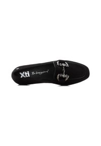 Zapato negro de ante tipo slip-on con suela plana, que presenta un diseño de hebilla metálica en el empeine y una plantilla texturizada con marca.