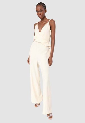 Donna con capelli intrecciati che indossa un top avvolgente senza maniche color crema e pantaloni ampi coordinati, in piedi davanti a uno sfondo semplice.