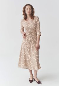 Beige gepunktetes Kleid mit kurzen Ärmeln und V-Ausschnitt. Verfügt über gathered Details an der Taille und einen ausgestellten Saum. Getragen mit burgunderfarbenen Absatzschuhen.
