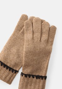 Et par beige strikkede handsker med sort zigzag-detalje nær de ribbede håndled på en hvid baggrund.
