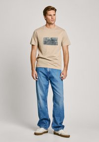 Bežinė medvilninė marškinė su stačiakampiu mėlynu ir pilku grafikų spaudiniu, kuriame užrašyta "Pepe Jeans", derinamos su šviesiai mėlynomis džinsų kelnėmis.