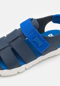 Tummansininen sandaali, joka koostuu tekstuurituista synteettisistä remmeistä ja pehmeästä sinisestä pohjasta, jossa on valkoinen kumipohja.