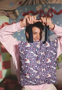 Mosolygó személy rózsaszín pulóverben virágmintás bevásárlótáskát tart, melyen őz, mókusok és nyulak láthatók, élénk színű háttér előtt.