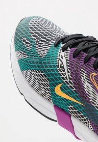 Färgglad sneaker med ett mesh-mönster i teal, svart och lila med en gul accent. Svarta snören, texturerad yta och vit sula.