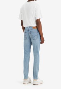 Personne portant un jean bleu clair, des baskets blanches et un t-shirt blanc uni, debout vue de dos sur un fond blanc.
