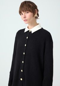 Young person with short brown hair wearing a black ribbed cardigan with gold buttons and a white collar against a plain light background.