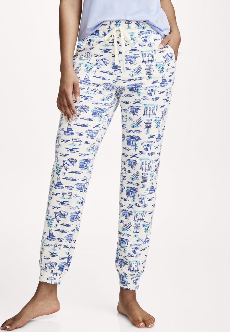 Vrouw draagt witte joggerbroek met blauwe illustraties van een meer, inclusief boten, borden en water, gecombineerd met een lichtblauw shirt.