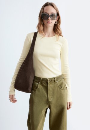 Giovane donna che indossa occhiali da sole oversize grigi, maglia crema a maniche lunghe, pantaloni a vita alta color oliva, con una grande borsa a tracolla marrone scuro.
