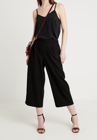 Camicetta di satin nera con spalline sottili, abbinata a culotte nere larghe. Il modello indossa sandali con tacco in stampa animale e una borsa a tracolla bordeaux.