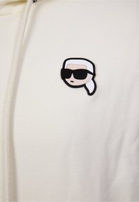 Cream hoodie featuring a black and white embroidered patch of a face with sunglasses. Material appears soft with a smooth texture.