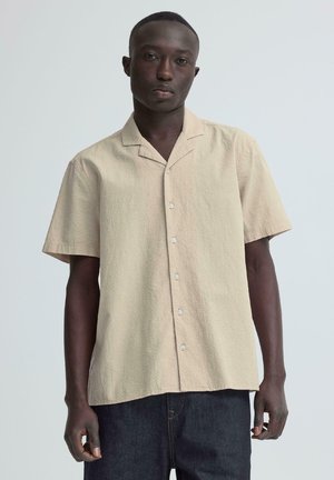 Mladi moški s temno poltjo, oblečen v bež kratko srajco z vzorcem z gumbi in temne hlače, stoji pred svetlo sivim ozadjem.