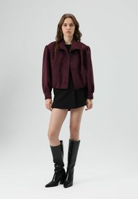 Bordeauxkleurig cropped jasje met pofmouwen en zwarte knopen, gecombineerd met een zwarte minirok en kniehoge zwarte laarzen, op een neutrale achtergrond.