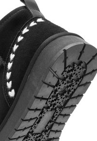 Zwarte suede schoen met witte stikseldetails, textuur rubberen zool met een sneeuwvlokpatroon, ontworpen voor tractie en duurzaamheid.