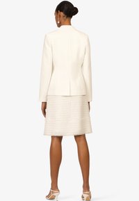 Blazer blanc ajusté associé à une jupe en crème texturée. La jupe présente un ourlet effiloché et des embellissements subtils, accompagnée de sandales à talons de couleur claire.