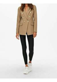 Beżowa dwurzędowa marynarka z dużymi srebrnymi guzikami, dwie kieszenie z przodu, w połączeniu z czarnymi legginsami i białymi trampkami.