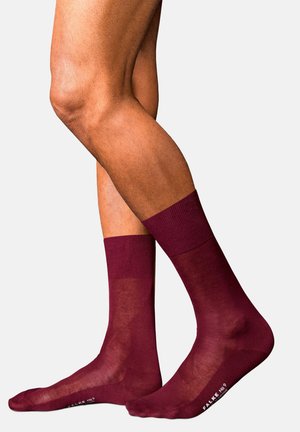 Piernas con calcetines transparentes burdeos hasta media pantorrilla con puños acanalados, mostrando piel suave y texto impreso cerca de los dedos sobre un fondo claro y liso.