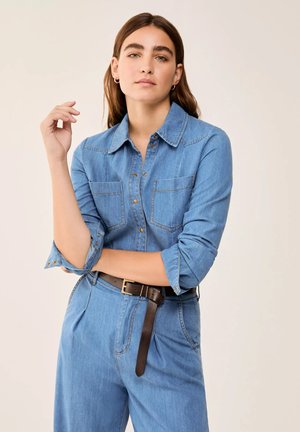 Giovane donna dai capelli lunghi e castani che indossa una camicia di jeans abbottonata e pantaloni di jeans abbinati con una cintura marrone, in piedi su uno sfondo beige.