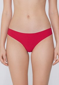 Eine Frau trägt schlichte rote Low-Rise-Bikini-Hosen, die den Mittelteil und die Oberschenkeloberseiten vor einem neutralen Hintergrund zeigen.