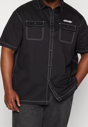 Uomo che indossa una camicia nera a maniche corte con bottoni e cuciture a contrasto bianche, con due tasche a patta frontali, mano sinistra in tasca.