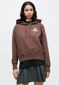 Καφέ φούτερ με κουκούλα και τσέπη τύπου ποδιά στο μπροστινό μέρος, με λευκό λογότυπο "Nike Athletics" και υφή φινιρίσματος. Φορεμένο με μαύρο εσωτερικό στρώμα και γκρι φούστα.