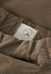 Brūns korduroja audums ar baltu sietu tagu un apaļu plastmasas pogu. Tā struktūra ir mīksta ar redzamām vertikālām rievām.