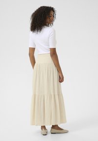 Light beige tiered maxi skirt with a smocked waistband, made of textured fabric. Paired with a white fitted top and beige flats.