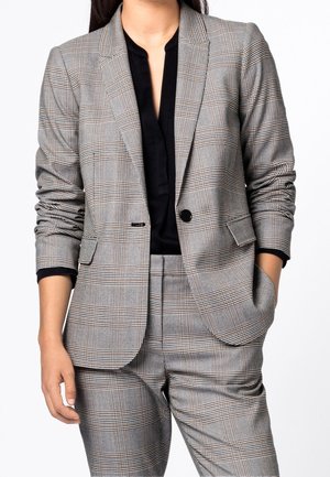 Persoon die een grijs geruit pak draagt met bijpassende blazer en broek over een zwart topje, staand met één hand in de zak tegen een witte achtergrond.