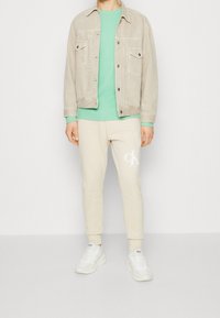 Beige Jeansjacke, mintgrünes Langarmshirt, cremefarbene Jogginghose mit Logo, weiße Sneakers. Lässiger Schnitt, weiche Materialien und schlichtes Design.