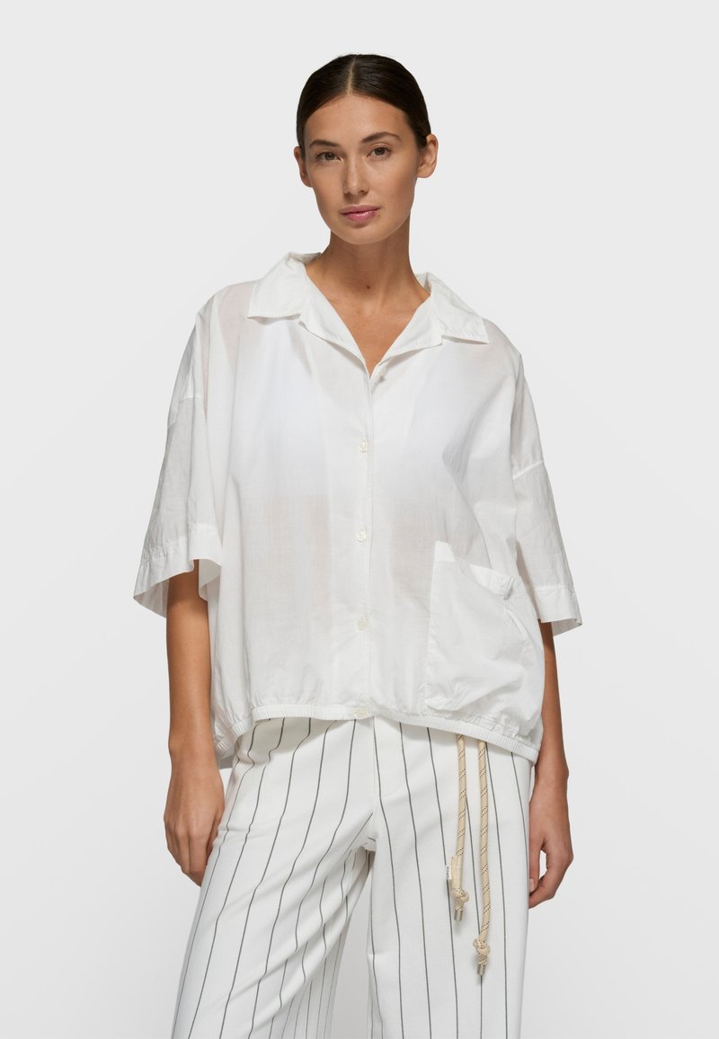 Chemise blanche à manches courtes avec boutons, offrant une coupe décontractée avec un ourlet tombant et deux poches avant. Tissu léger et transparent avec une texture subtile.