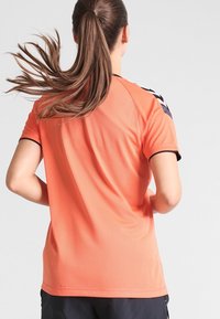 Tričko s krátkým rukávem sportovního typu v jasně oranžové barvě s texturovanou, prodyšnou tkaninou. Černé akcenty na rukávech mají vzor ve tvaru šípů.