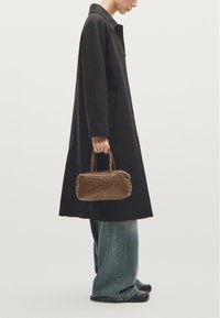 Geflochtener brauner Handtasche mit runder Form und kurzen Griffen, gehalten von einer Person, die einen schwarzen Mantel und weite blaue Jeans trägt.