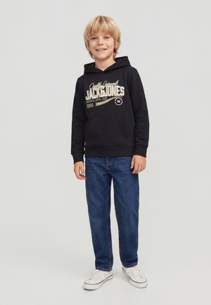Uśmiechnięty blond chłopiec stoi, ubrany w czarną bluzę z kapturem Jack & Jones, niebieskie dżinsy i białe trampki na jednolitym jasnoszarym tle.