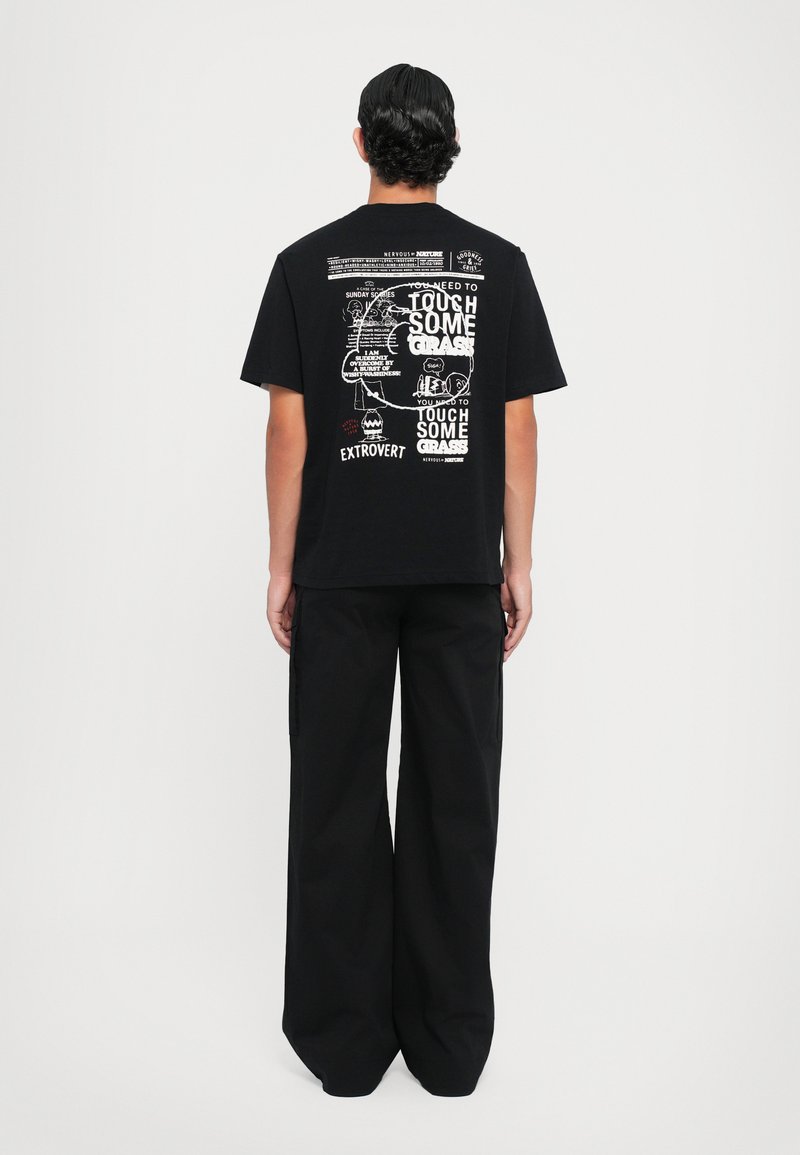 Schwarz Baumwoll-T-Shirt mit einem grafischen Design auf dem Rücken, das weiße Texte und Zeichnungen zeigt. Kombiniert mit lässigen, locker sitzenden schwarzen Hosen.