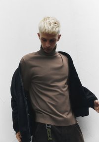 Junger Mann mit kurzen blonden Haaren, der einen braunen Rollkragenpullover trägt und eine schwarze Jacke vor einem weißen strukturierten Wandhintergrund hält.